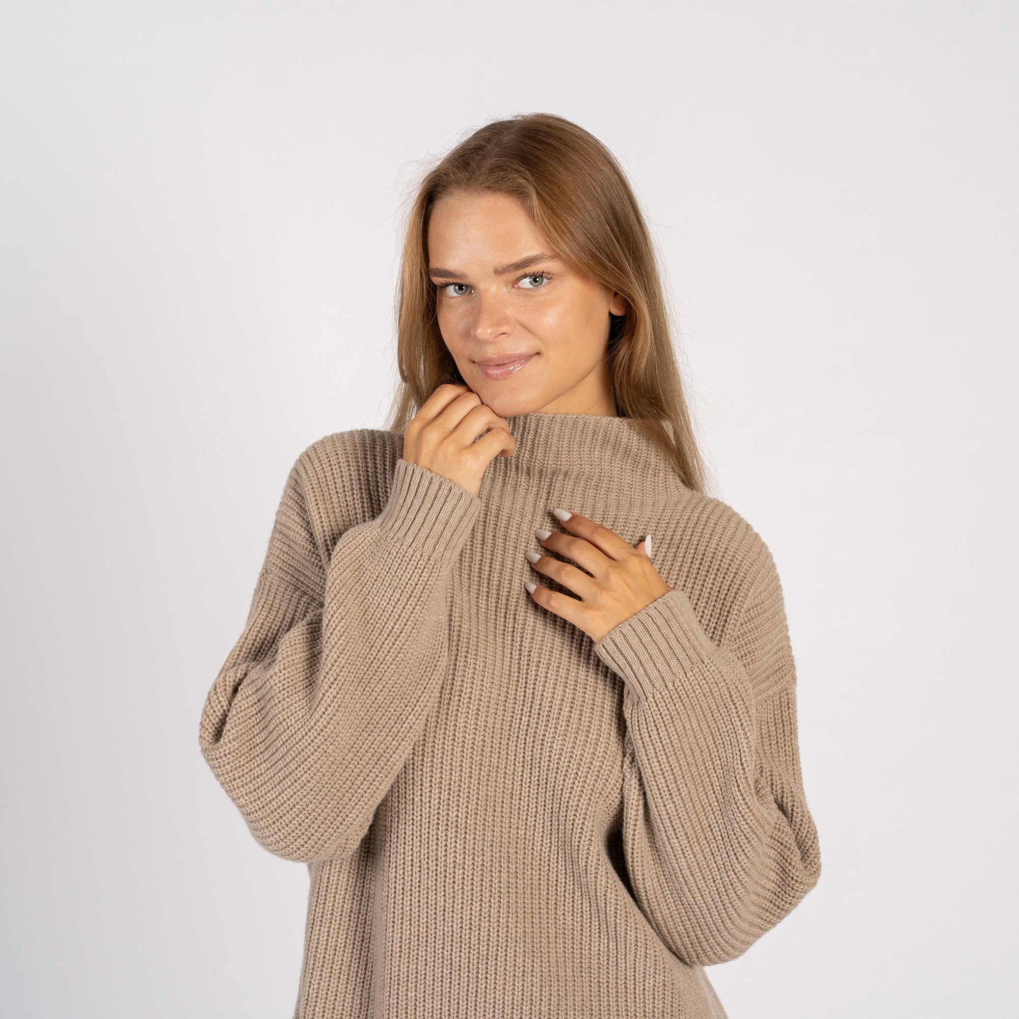 A woman smiling while wearing a loose beige knit turtleneck sweater with side slits, paired with light blue jeans.