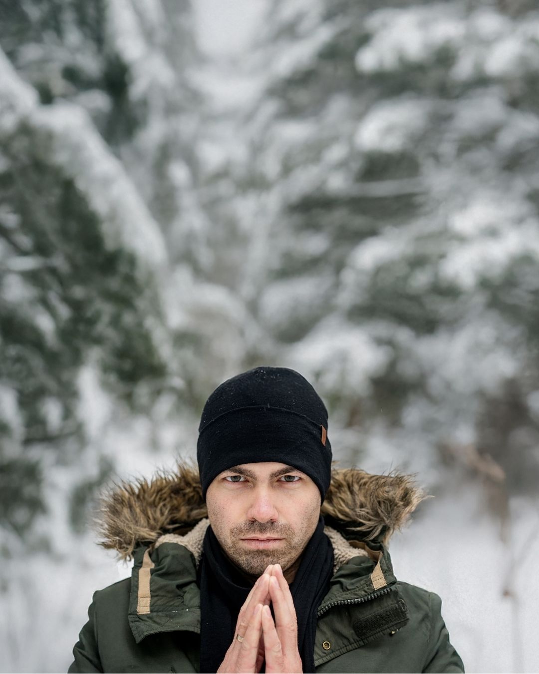 Man wearing winter coat and black beanie outdoors with snow-covered trees in the background