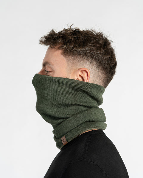 A man with short, dark, wavy hair, wearing a dark green knit neck gaiter pulled up to cover his nose and mouth, and a black long-sleeved shirt. He is shown in profile, facing right with his eyes closed. A small brown leather tag is visible on the lower front of the gaiter. The background is plain white.