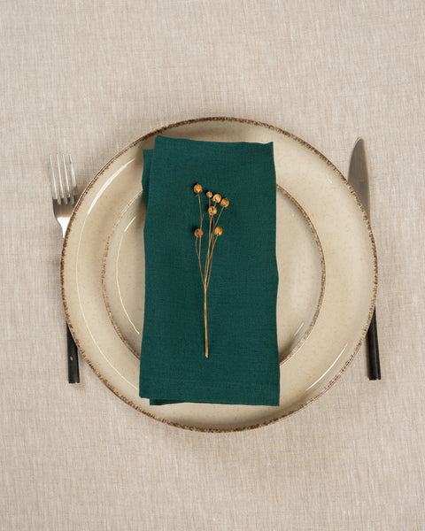 The menique linen napkin in dark green color, put on top of two stacked plates . There are cutlery items by the sides of the plates. The background is a natural color linen tablecloth.