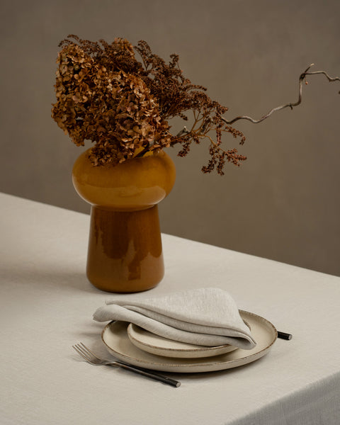 A linen tablecloth covering  tabletop. On top of the table is a vase and two stacked plates with cutlery by the sides. The plates have a natural color linen napkin on top of them folded into a neat triangle.