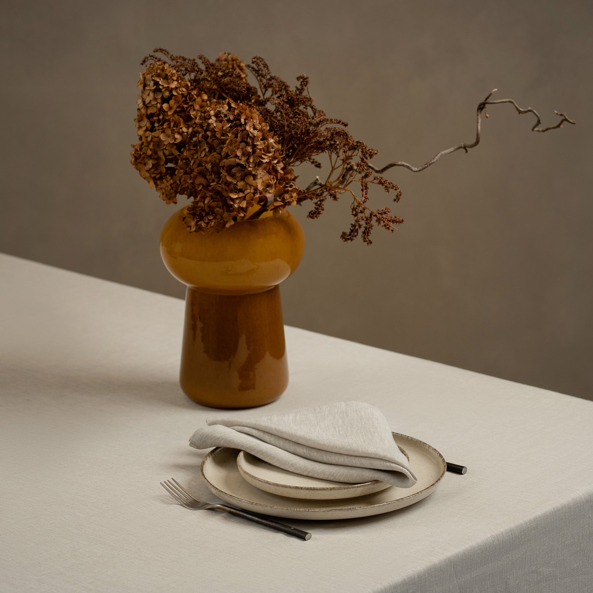 A tabletop covered with a linen tablecloth, with a vase and a single plate set on top of it. By the sides of the plates there are cutlery items, and there is a linen napkin in cobalt blue color on top.