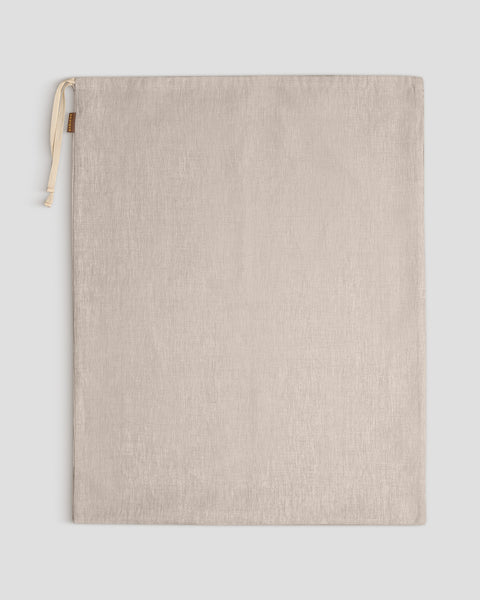 A large natural color linen organizing bag lies flat against a light gray background, its light-colored drawstrings gathered at the top left, next to a small brown tag.