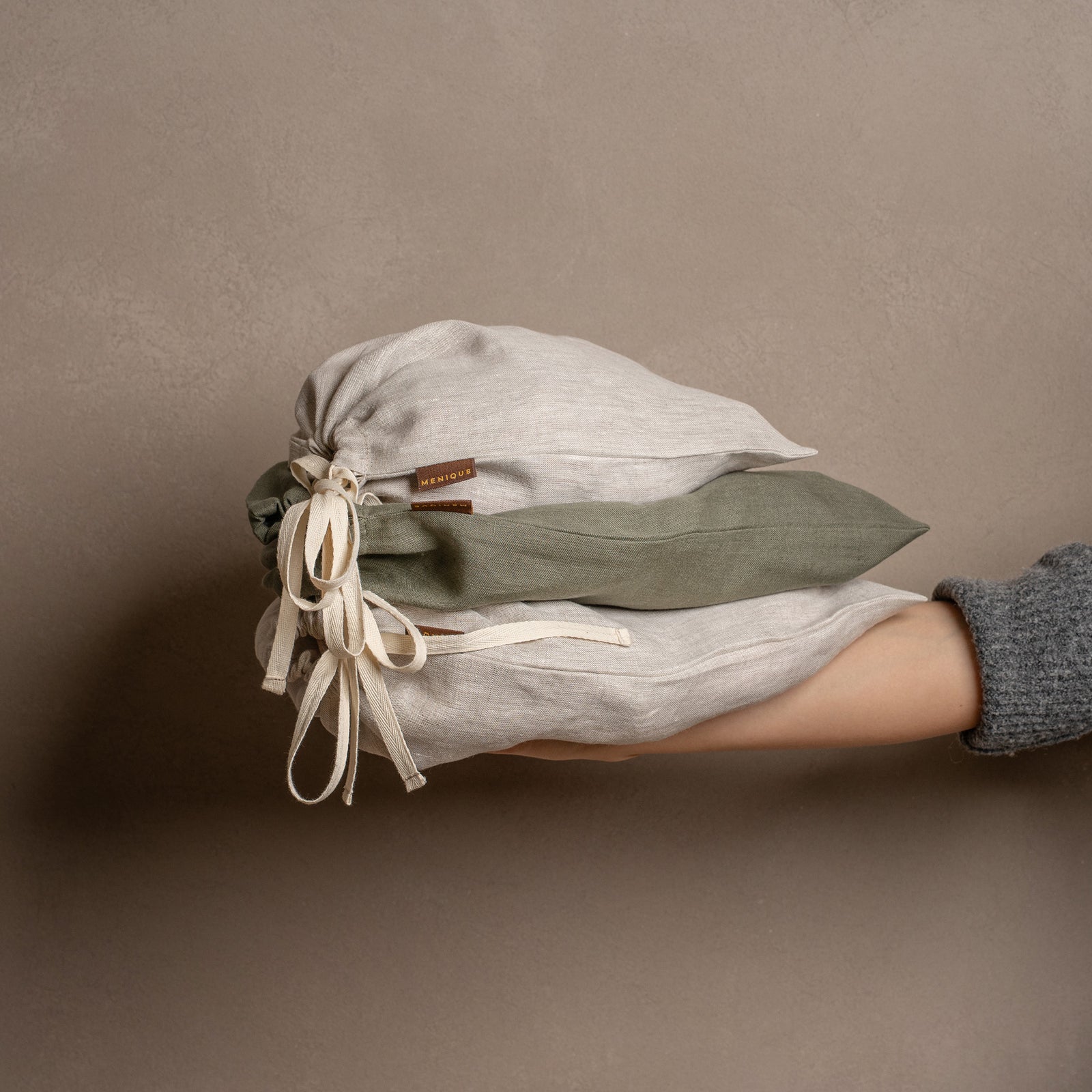 Five closed linen drawstring bags in varying sizes and colors of natural and stone green are arranged on a textured jute rug against a warm brown wall.