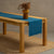 A wooden table positioned on top of a carpet. There is a dark green linen table runner and a vase on top.