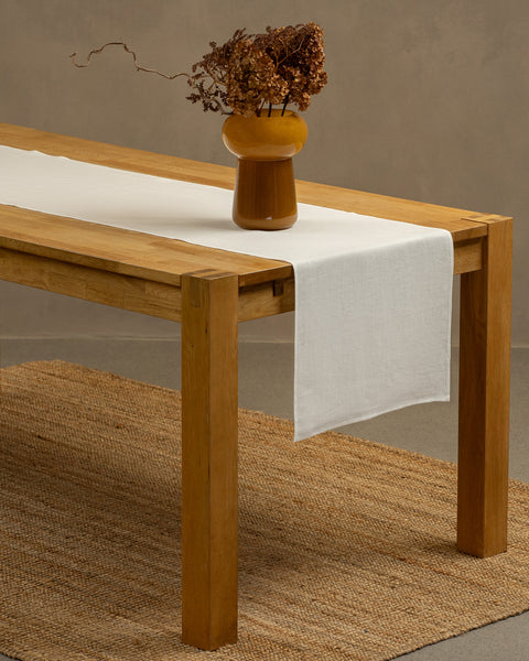 A wooden table positioned on top of a carpet. There is a pure white linen table runner and a vase on top.