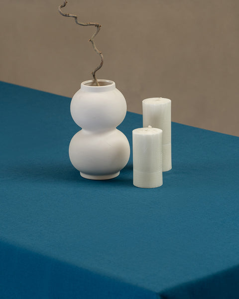 The menique linen tablecloth in cobalt blue color, with a modern white vase on top of it and two different height white candles by the sides.