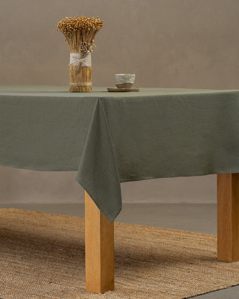 The menique linen tablecloth in stone green color, draping over a wooden table, with a vase on top of it, and a clay cup and plate by the side.