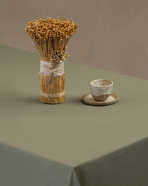 The menique linen tablecloth in stone green color, draping over a wooden table, with a vase on top of it, and a clay cup and plate by the side.