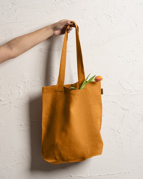A fair-skinned arm and hand hold up a spicy yellow durable linen tote bag against a textured off-white wall. A single tulip with a red and yellow bloom and green leaves is visible inside the bag.
