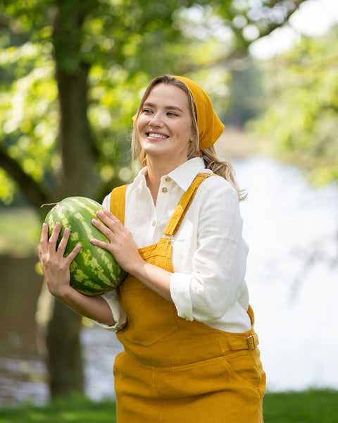 The same woman in the mustard yellow outfit stands in a sunlit park with trees and a body of water in the background. She is smiling brightly while holding a large, whole watermelon in her arms.