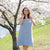 A woman wearing a cloudy blue linen slip dress stands beside a tree covered in white blossoms and gently smells a cluster of flowers.