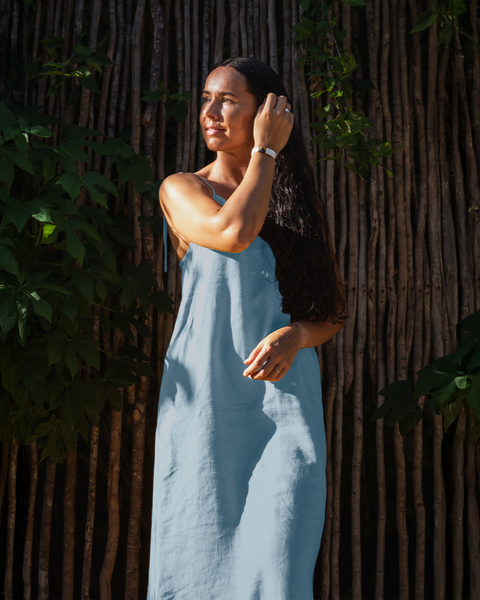 A person with long, dark hair and light skin is standing outdoors, looking towards the left with a hand near their hair. They are wearing a cloudy blue sleeveless dress with thin straps. The background features a textured wooden fence or wall partially obscured by green foliage.