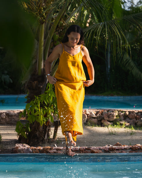A woman with dark hair wearing a yellow linen sundress. Behind her is a bright blue pool surrounded by palm trees and tropical foliage. The ground is light-colored with shadows.
