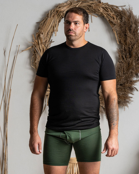 A man standing indoors, looking off to the left with a neutral expression. He is wearing a fitted black short-sleeved t-shirt and olive green boxer briefs. He has a tattoo on his left forearm. Behind him is a white wall decorated with a large, rustic circular wreath made of dried grasses and reeds.
