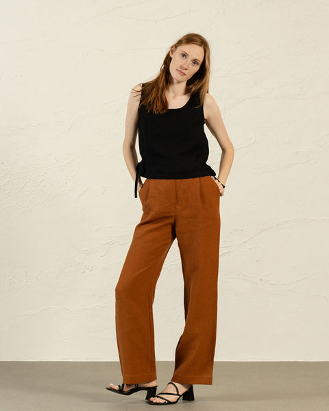 A full-body studio shot of a woman with long, light brown hair standing with her hands in her pockets and looking directly at the camera. She is wearing a black sleeveless top with a scoop neck and side ties, paired with rust-colored, wide-leg trousers. She is also wearing black open-toed sandals with a block heel. The background is a plain, light-colored wall.