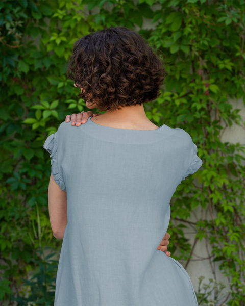 A woman with short, dark curly hair is seen from the back, wearing a cloudy blue linen dress. She is standing in front of a wall covered with green ivy, and her arms are crossed with her hands resting on her shoulders.