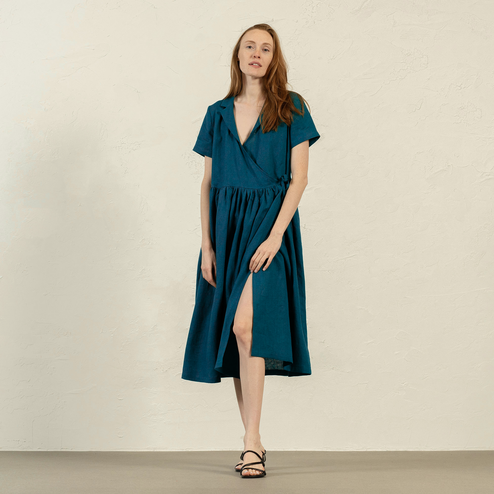 A woman with brown hair, wearing a light blue, short-sleeved linen wrap dress, stands outdoors next to a tree with white blossoms.
