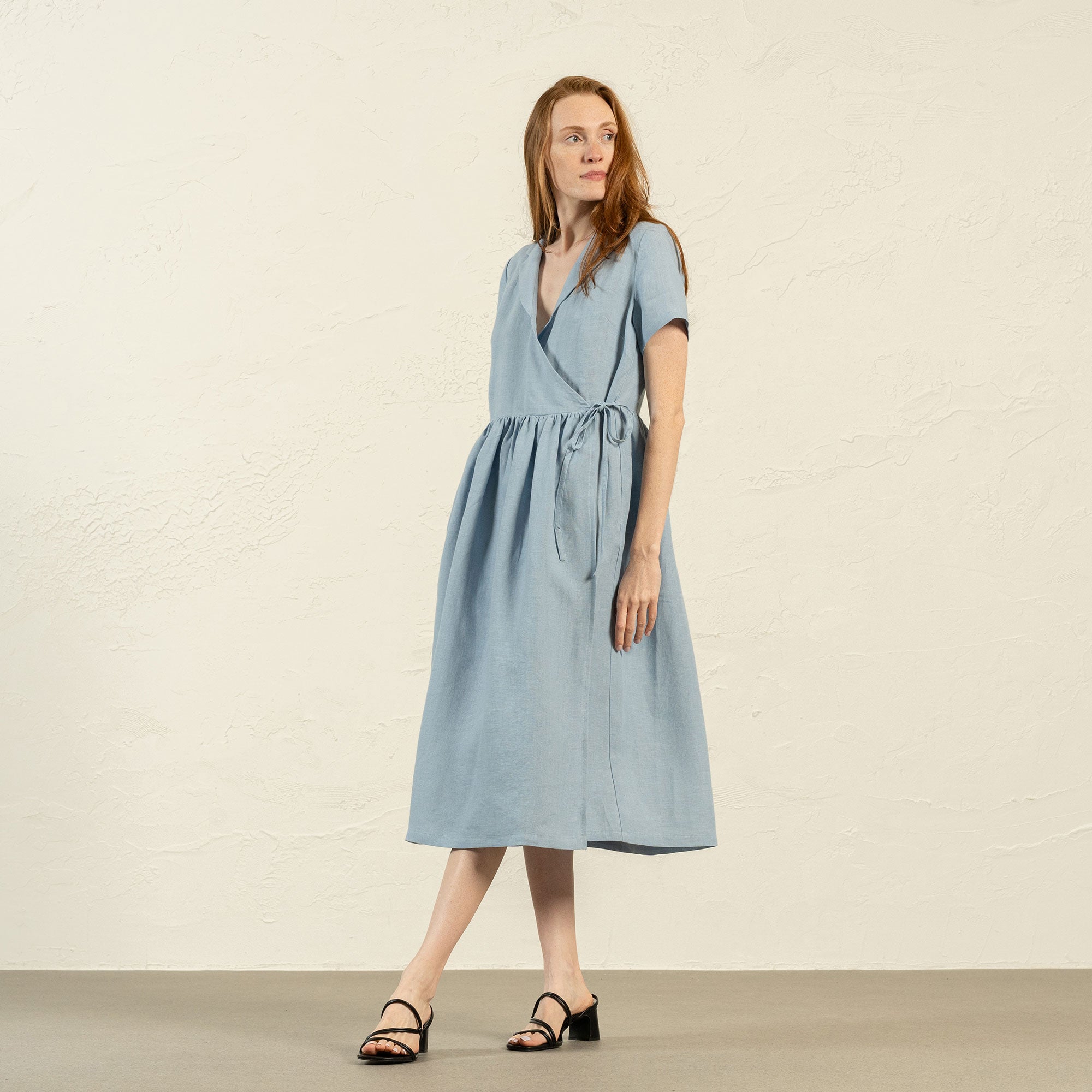A woman with brown hair, wearing a light blue, short-sleeved linen wrap dress, stands outdoors next to a tree with white blossoms.
