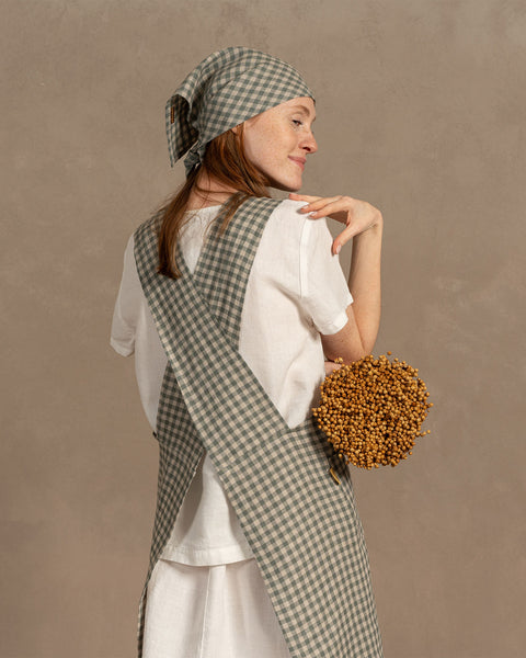 A person wearing a green gingham cross-back linen apron and matching headscarf, shown from behind, holding a round dried flower bouquet against a neutral beige backdrop.