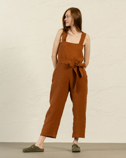A person with long reddish-brown hair and light skin is standing at a slight angle, looking to the left. They are wearing pinafore linen jumpsuit with wide straps and a tied belt, paired with olive-green slip-on shoes.