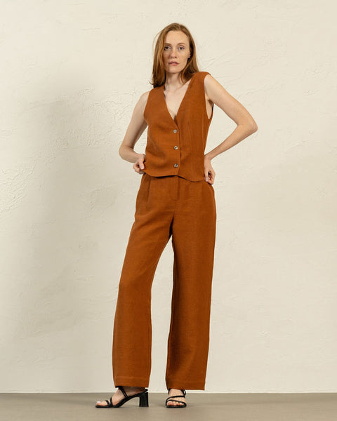 A fair-skinned woman with shoulder-length brown hair stands facing forward, modeling an almond brown linen vest and menique wide-legged trousers. She has her hands resting on her hips and is wearing black open-toe sandals with a low heel. The background is a textured light beige wall and a light gray floor.