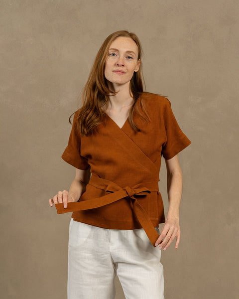 A fair-skinned person with long, auburn hair, wearing V-neck linen wrap top with short, wide sleeves and light-colored pants. They are holding the ends of the top&#39;s tie in front of them, forming a bow.