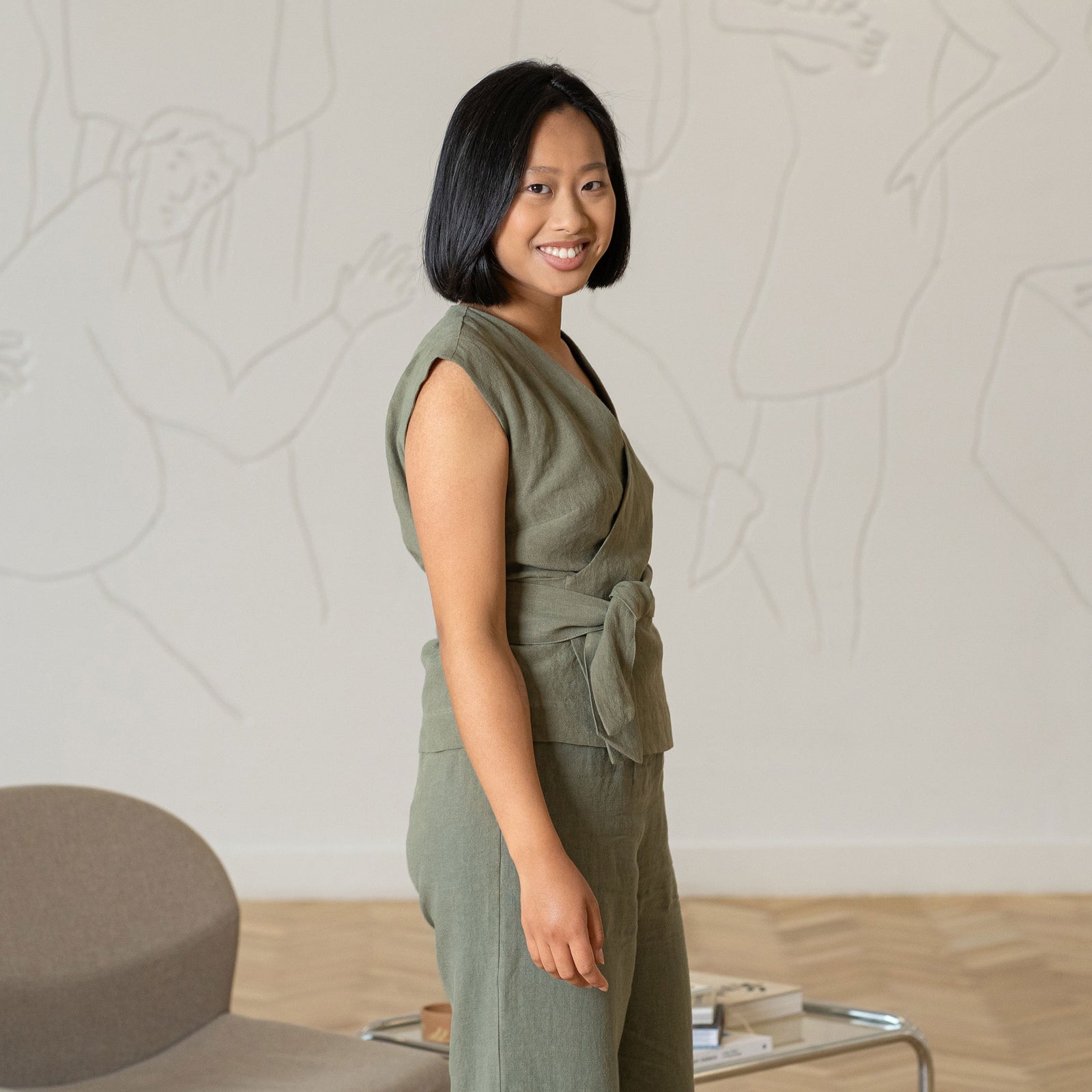 A young woman with a short, dark bob haircut stands facing forward in an indoor setting. She is wearing a stone green linen two-piece outfit consisting of a sleeveless V-neck wrap top with a tie waist and wide-leg trousers. Her hands are clasped in front of her. She is wearing dark open-toed sandals.