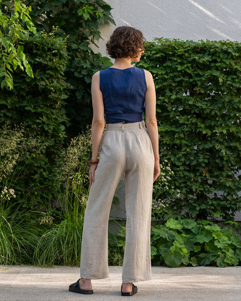 From the back, a light-skinned person with short, wavy brown hair is standing on a paved surface surrounded by greenery. They are wearing a storm blue sleeveless wrap top and natural wide-legged pants, along with dark slip-on shoes.
