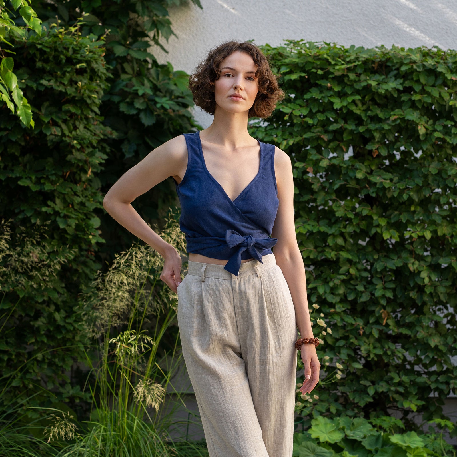A fair-skinned person with long reddish-brown hair and freckles is standing outdoors with a slight smile. They are wearing a pure white linen cropped wrap top tied at the side with a wide cream-colored ribbon, and stone green pants. The background includes trees, a black metal fence, and a light-colored building.