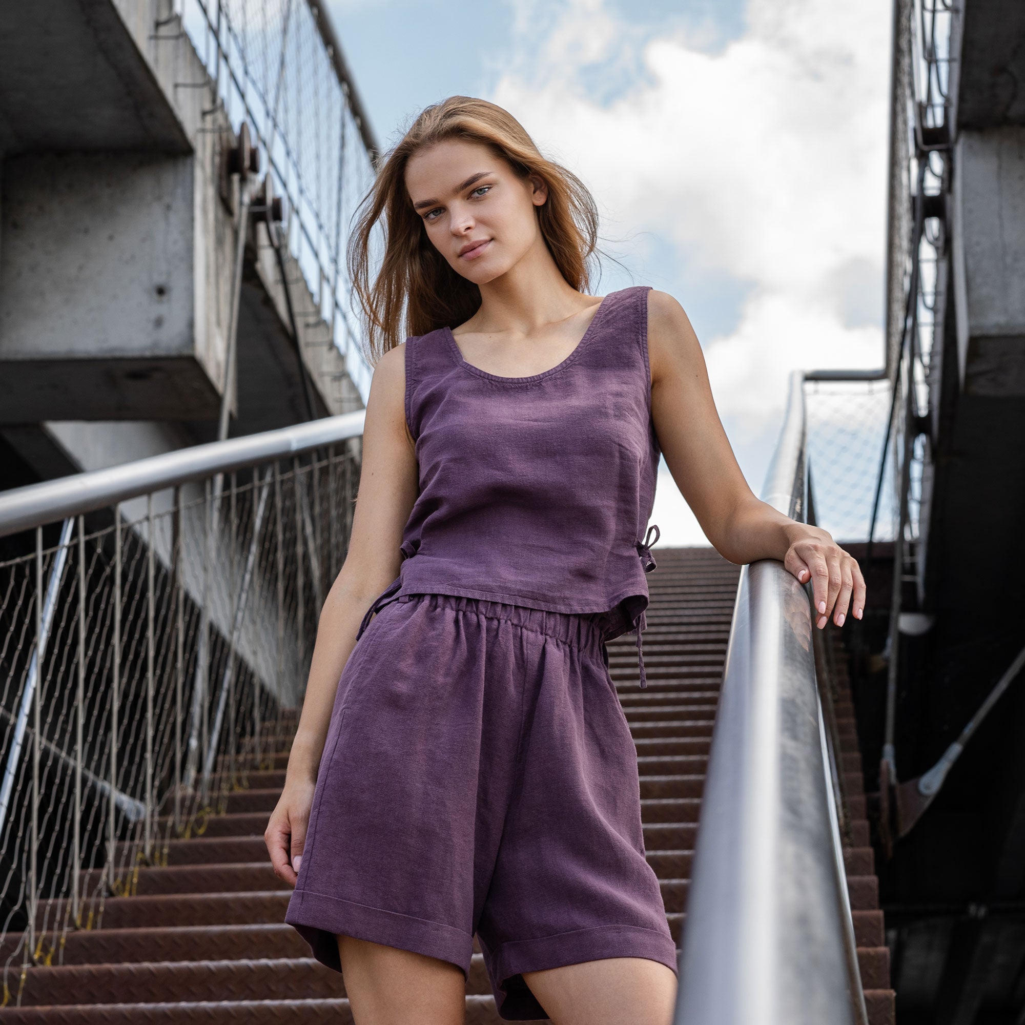 A woman with blonde hair pulled back wears a natural linen sleeveless crop top with side ties, paired with matching light natural linen pants. She stands facing forward with one hand in her pocket against a muted brown textured background.
