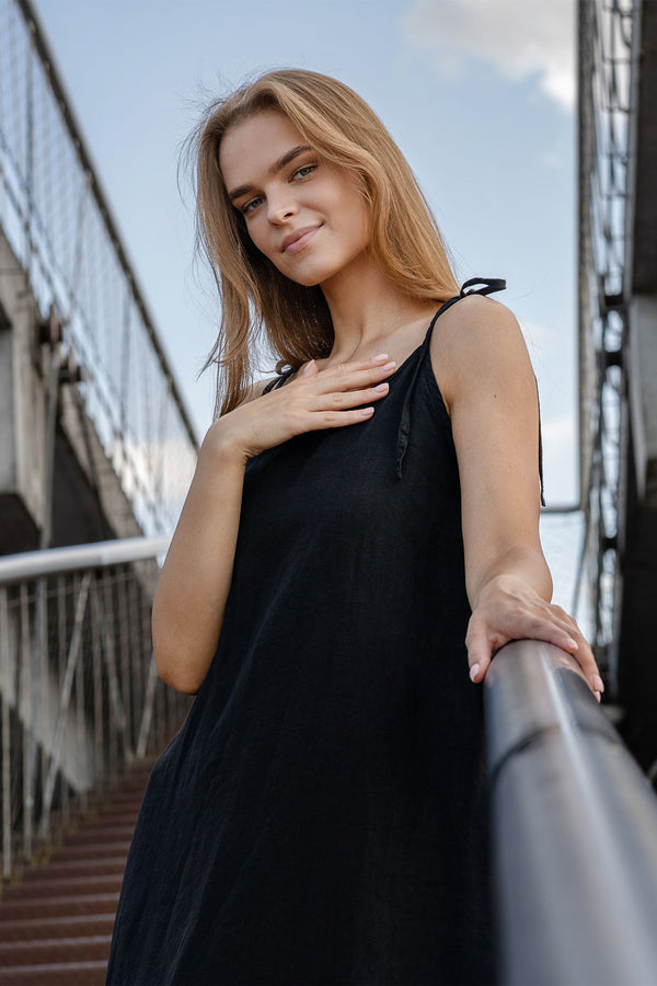 A light-skinned person with long blonde hair is standing on a set of outdoor stairs, looking towards the camera with a gentle smile. They are wearing a pure black sleeveless linen dress with thin straps, and one hand is resting on their chest while the other holds onto a metal railing. The background shows a cloudy sky and architectural elements of a walkway or bridge.