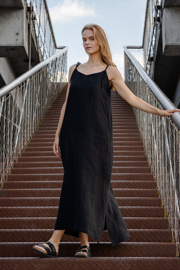 A light-skinned person with long blonde hair is standing on a set of metal stairs with a walkway above. They are wearing a long black menique sleeveless dress with thin straps and dark sandals, and are holding onto a metal railing with one hand. The background shows a somewhat overcast sky.