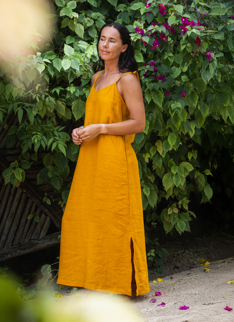 Woman wearing a mustard yellow linen maxi dress standing in a lush garden with green foliage and flowers.