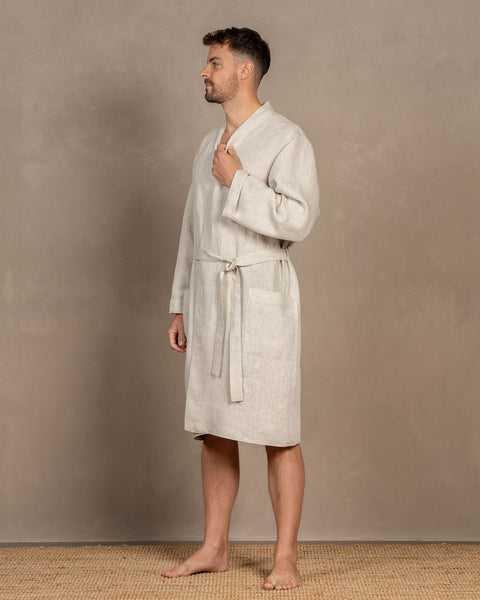 A man in a menique natural color linen robe stands in profile on a woven mat, with a textured grey wall behind him.