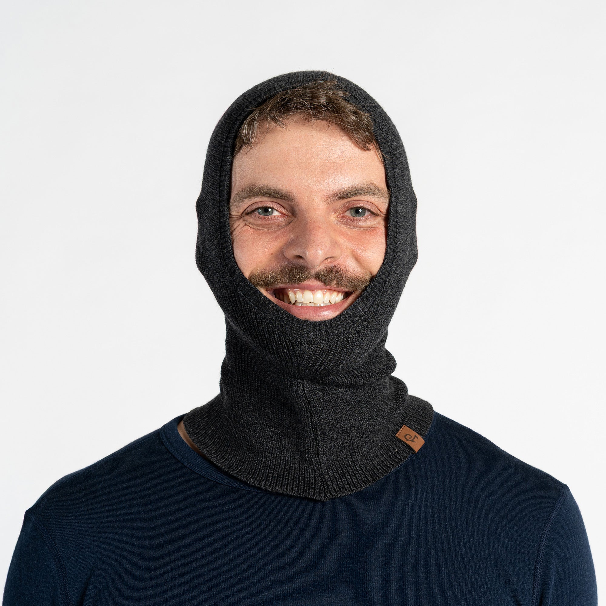 A man with a beard and mustache wears a dark green, fitted menique balaclava that covers his head and neck, with an opening for his face. He is also wearing a black long-sleeved shirt and is looking directly at the viewer with a smile.