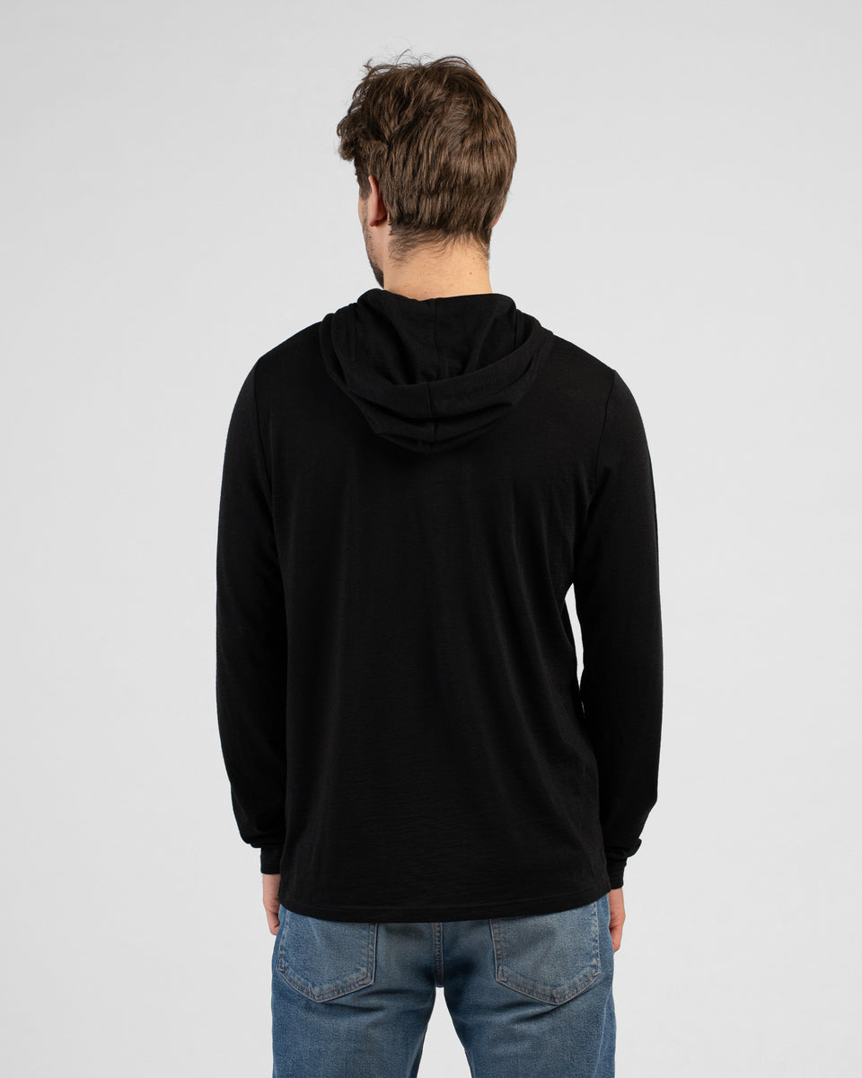 A studio shot showing the back of a man wearing a black long-sleeved hooded shirt with the hood down and blue jeans. He stands facing away from the camera against a plain light grey background.