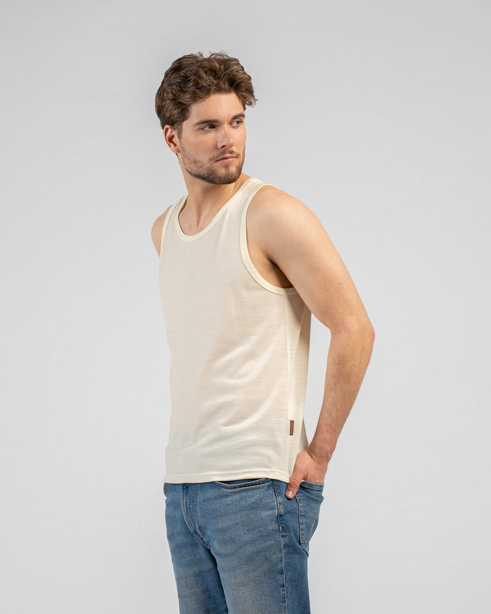 A studio portrait of a man standing and looking back over his right shoulder. He is wearing a light beige tank top and medium-wash blue jeans, with his left hand resting in his front pocket. The background is a solid light grey.