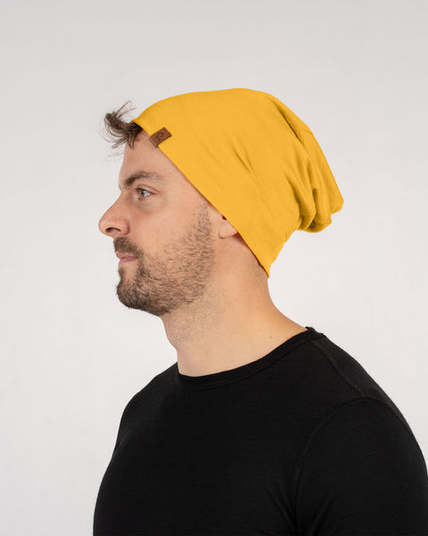 A man with short brown hair and a beard is shown in a profile view, wearing a spicy yellow merino wool beanie and a black t-shirt. He appears to be looking off to the left. The background is plain white.