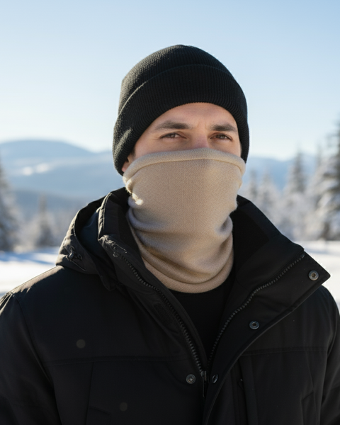 A man looks at the camera while wearing a black winter jacket, a black beanie, and a creamy beige knit neck gaiter that is pulled up over his nose and mouth. He is standing in a sunny, snow-covered landscape with evergreen trees and mountains in the background.
