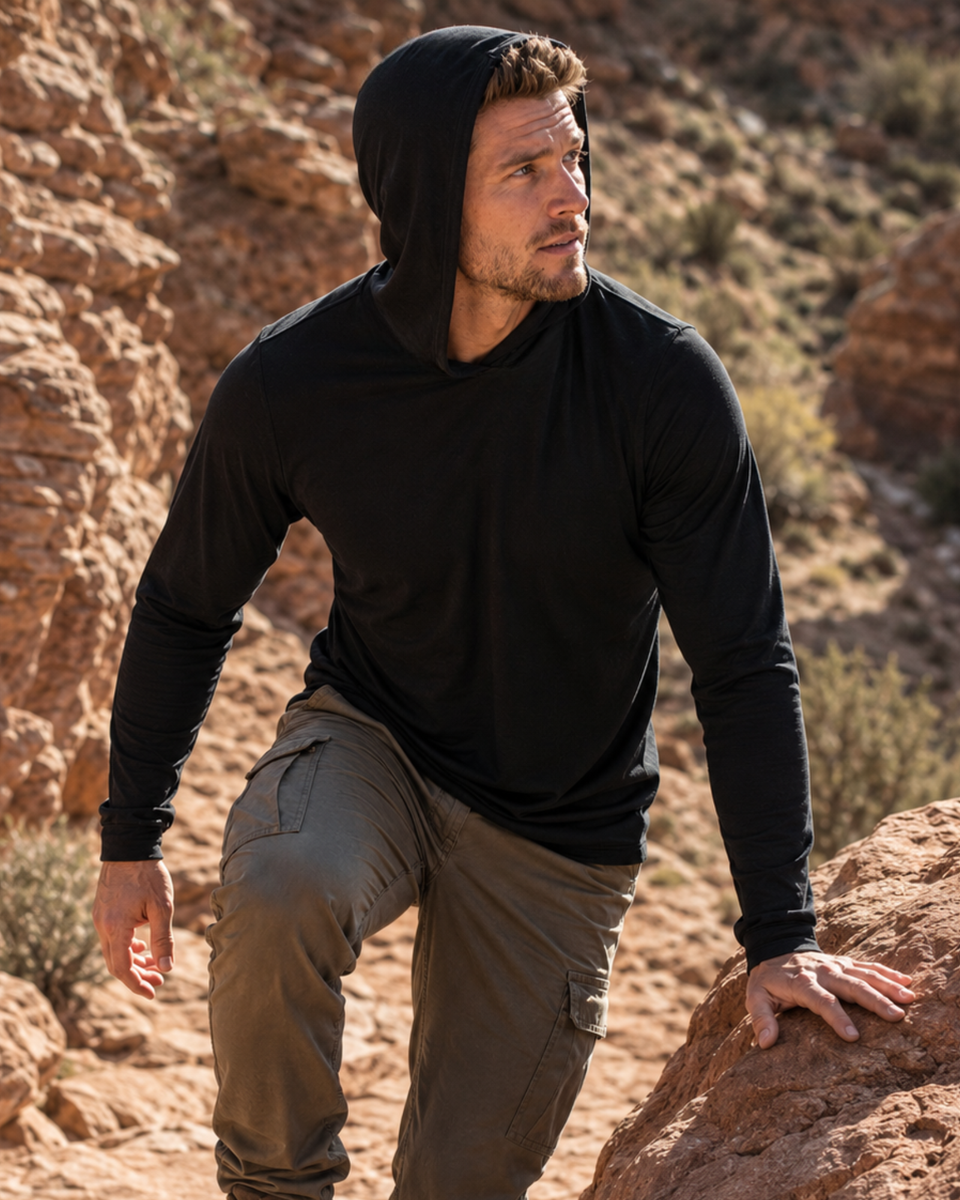 An outdoor shot of a man hiking along a rugged, sunlit rocky trail. He is looking off to the left, wearing a black long-sleeved hooded shirt with the hood down and olive green cargo pants.
