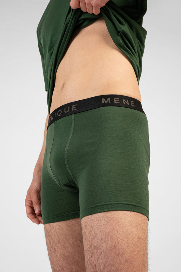 A close-up shot of a man's torso and thighs wearing dark green short boxer briefs with a black logo waistband. He is pulling up his matching dark green t-shirt to reveal his midriff against a light grey background.