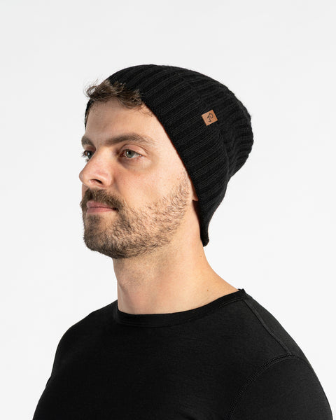 A person with light skin, short dark hair, a mustache, and light-colored eyes is shown in profile against a white background. They are wearing a black knit beanie with a small brown tag on the side and a black long-sleeved shirt.