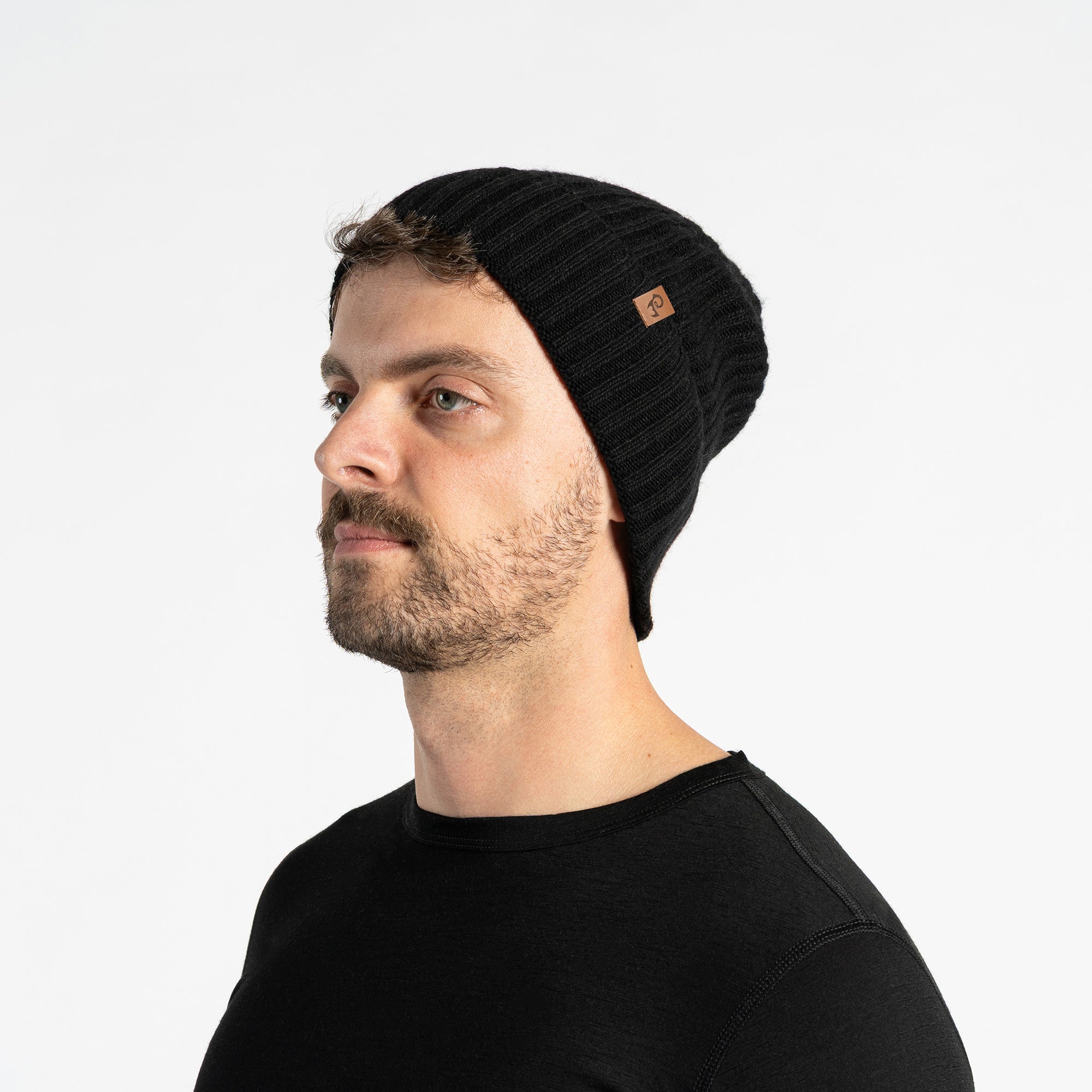 A person with light skin is shown in profile, smiling and looking to the left. They are wearing a black ribbed knit beanie with a small brown tag, a black turtleneck sweater, a black knit neck gaiter with a similar brown tag, and a black puffer jacket. The background appears to be concrete steps in an outdoor setting.