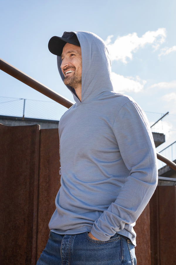 A smiling man standing outdoors near rusted metal barriers, wearing a dusty blue hoodie with the hood pulled up over a black baseball cap. He has one hand in his jeans pocket and is looking to the side.