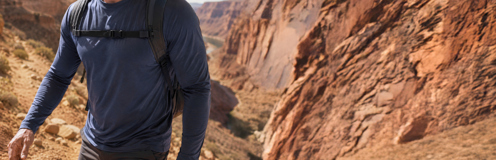 A man wearing a navy blue long-sleeve shirt and a black backpack hiking along a trail with a vast, deep red rock canyon in the background.