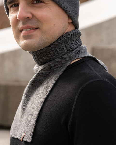 A close-up shot of a smiling man, showing his lower face and chest. He is wearing a dark grey knit beanie and a matching dark grey knit neck warmer with a ribbed collar and a flat front bib over a black long-sleeve shirt. A small brown logo patch is visible on the bottom corner of the neck warmer's bib. Blurred concrete stairs are in the background.