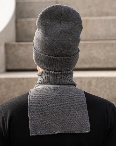 The back view of a man wearing a dark grey knit beanie and a matching dark grey knit neck warmer over a black long-sleeve shirt. The rectangular back panel of the neck warmer lies flat against his upper back. Blurred concrete stairs are in the background.