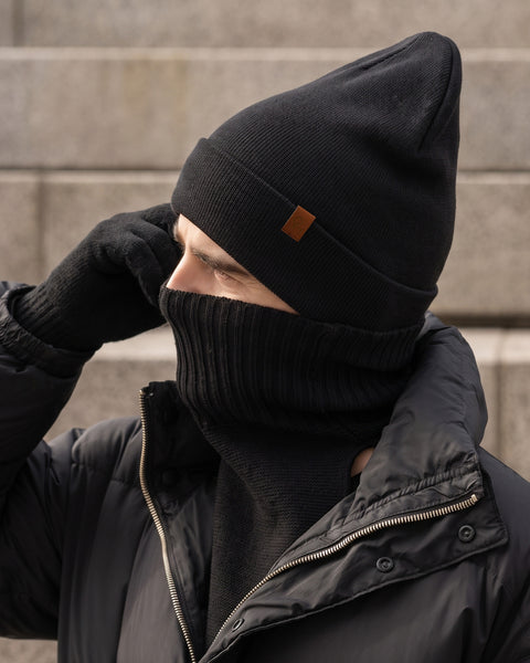 A man in profile looking to the left, dressed for cold weather. He is wearing a black winter jacket, black knit gloves, a black knit beanie with a small brown logo patch, and a black knit neck warmer pulled up to cover his nose and lower face. Out-of-focus concrete stairs form the background.