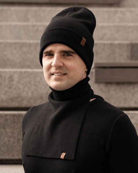 A smiling man facing the camera, wearing a black knit beanie with a small brown logo patch and a matching black knit neck warmer over a black long-sleeve shirt. The neck warmer features a ribbed turtleneck collar and a flat bib resting on his chest with a small brown logo tag at the bottom. Blurred concrete stairs are visible in the background.